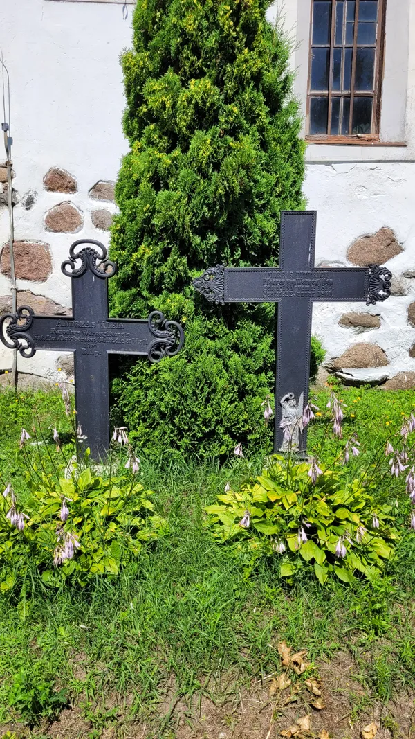 Dwa żeliwne, neogotyckie krzyże nagrobne dawnych, niemieckich mieszkańców wsi, oparte o żywopłot przy murze kościoła.